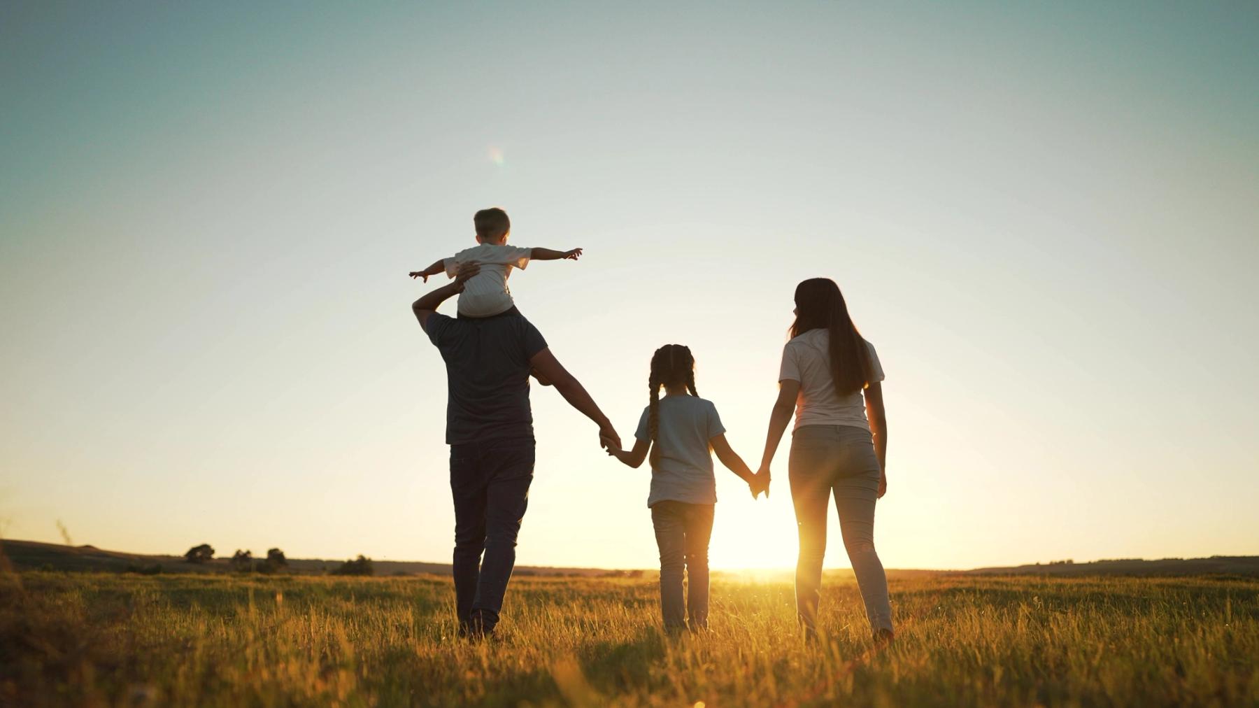 Image Alt Silhouetted family of four walking through a grassy field at sunset, with a child on the father's shoulders.