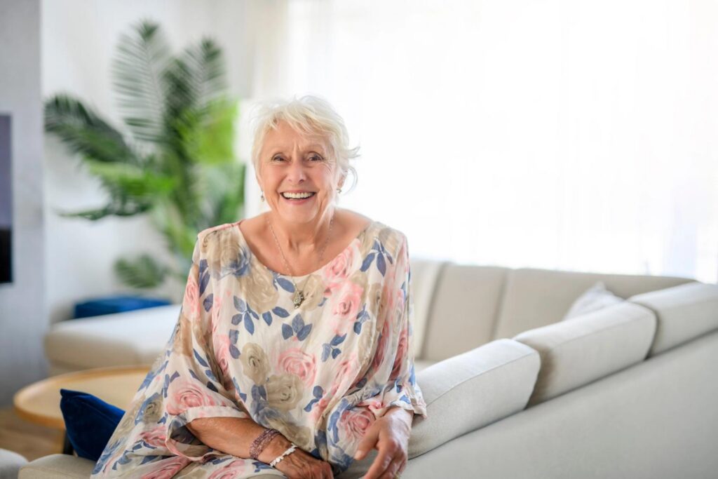 A smiling older woman with white hair wearing a floral top leans on a light-colored sofa.