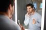 A man looking in a mirror holds a toothbrush and touches his cheek with a pained expression.