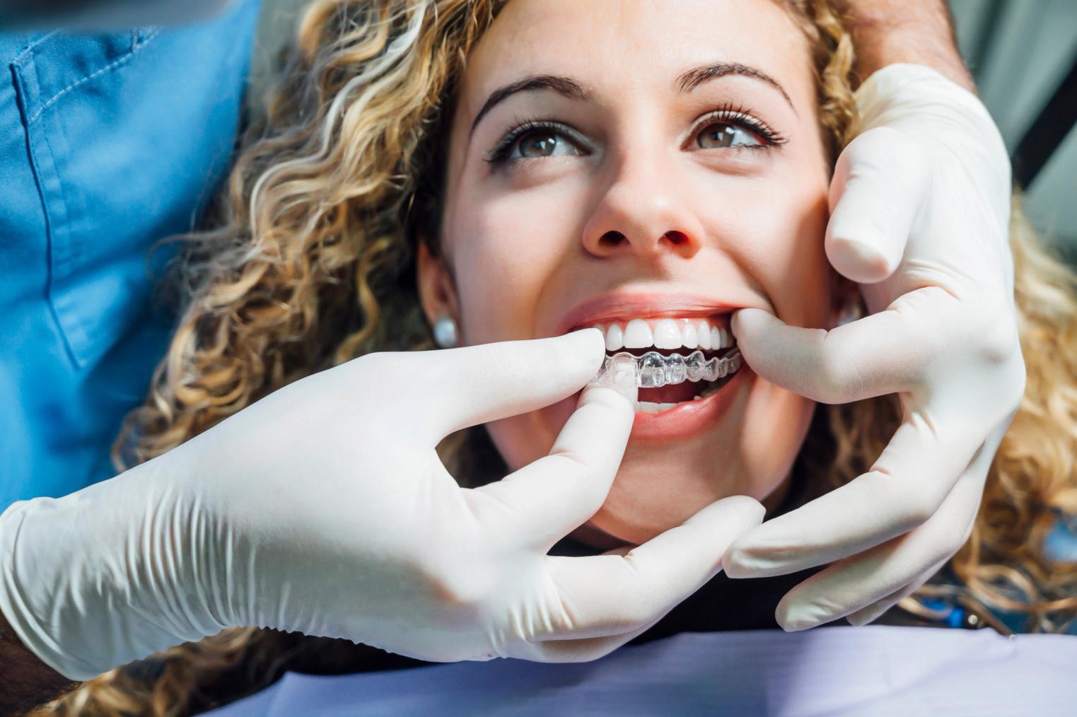 A smiling woman having clear aligners fitted by gloved hands.