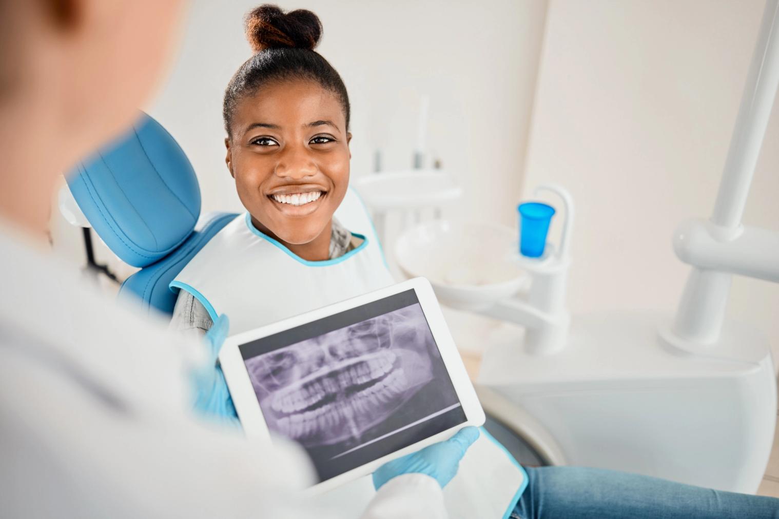 Image Alt Smiling young woman in a dental chair looking at an X-ray on a tablet held by a dentist.