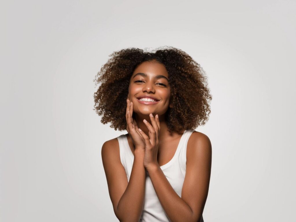 A smiling woman with curly hair gently touches her face.