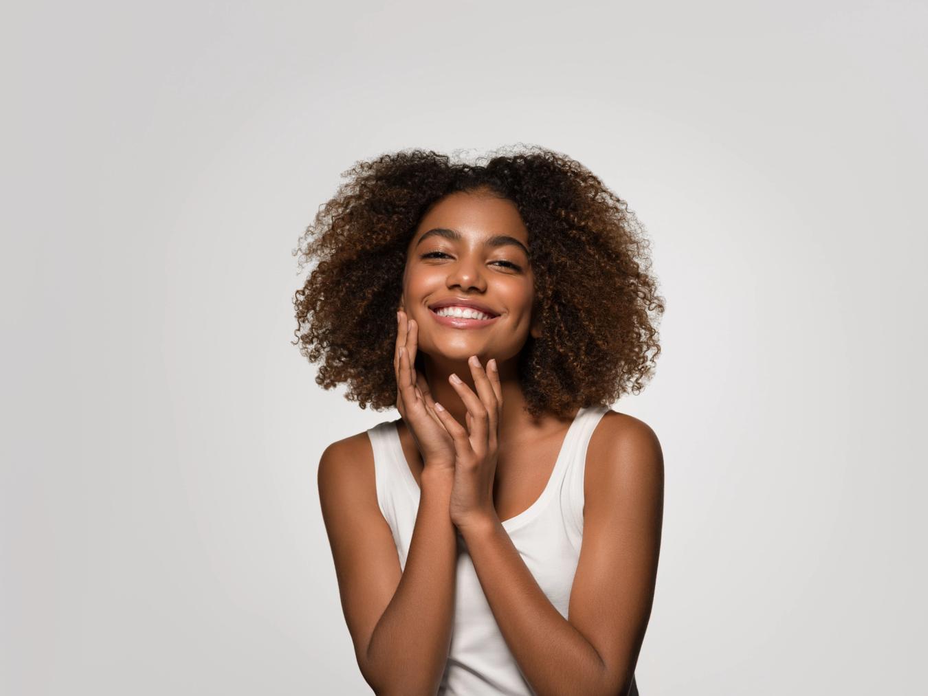 A smiling woman with curly hair gently touches her face.