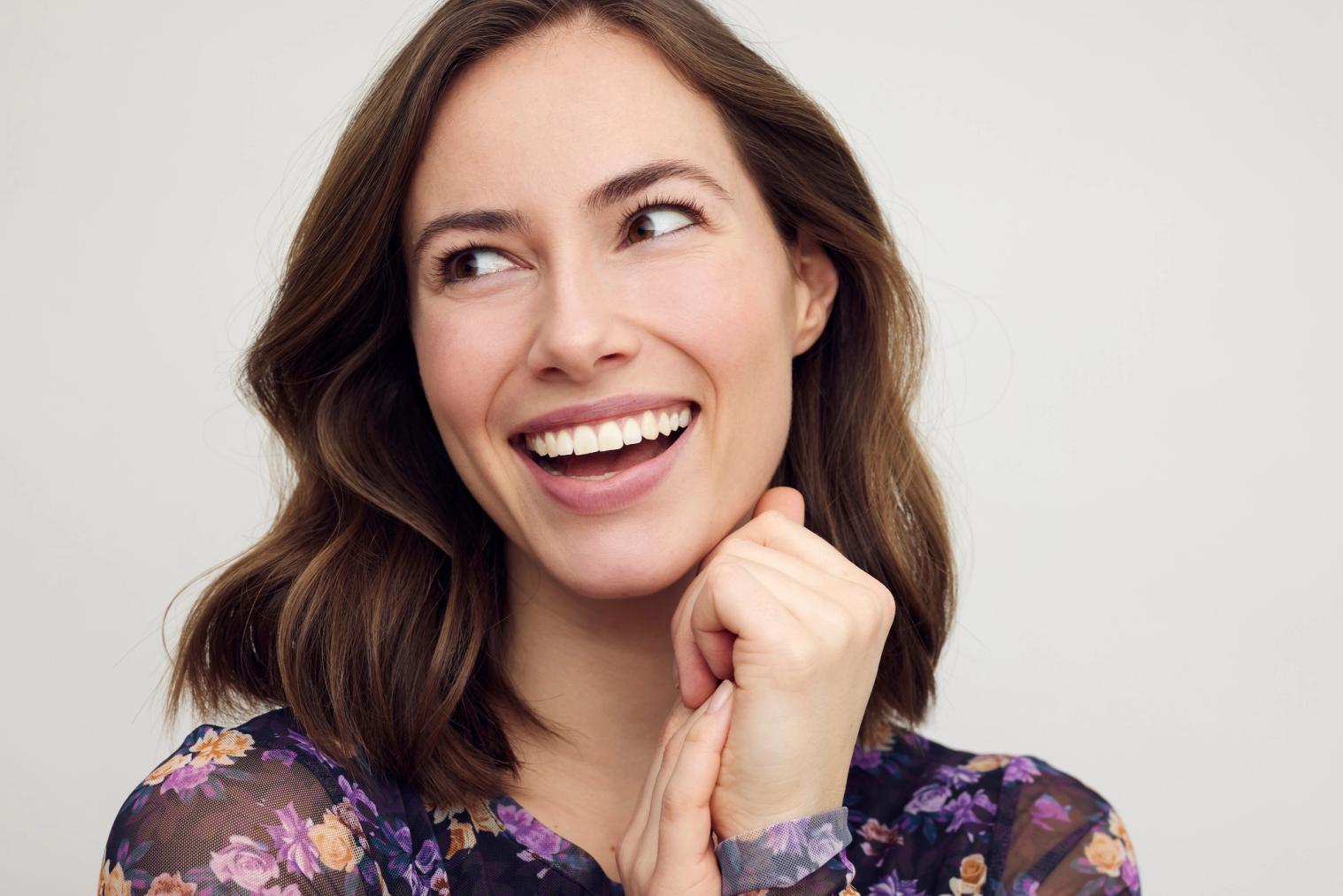 Image Alt A smiling woman with brown hair looks right, hands clasped under her chin.