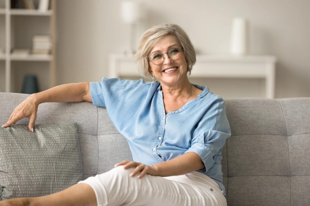 Smiling blonde woman with glasses sitting on a sofa.