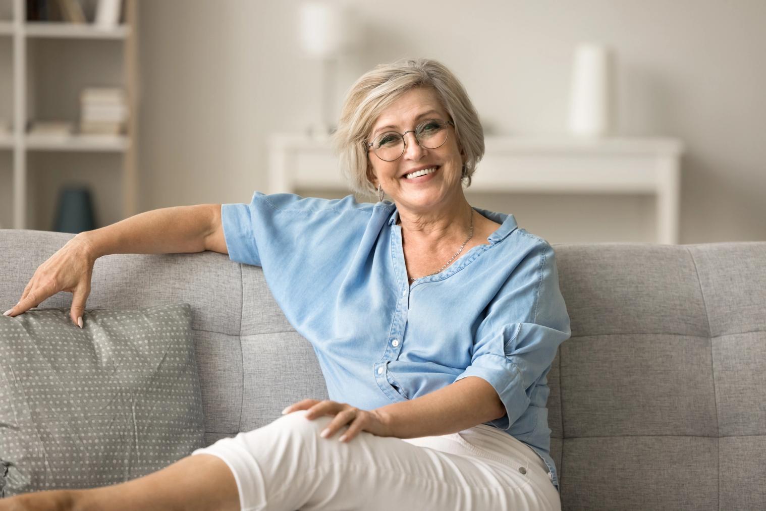 Image Alt Smiling blonde woman with glasses sitting on a sofa.